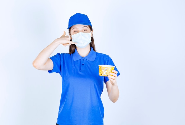 Young woman in uniform and medical mask with plastic cup making phone call gesture