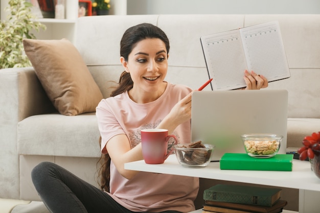 Young woman used laptop holding and points with pen at notebook sitting on floor behind coffee table in living room