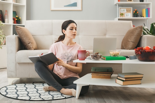 Young woman used laptop writes on notebook sitting on floor behind coffee table in living room