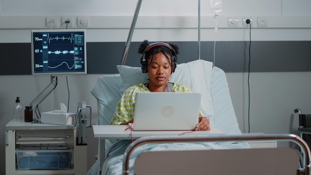 헤드폰과 노트북을 사용하는 젊은 여성은 질병을 치료하기 위해 기다리는 동안 병원 병동에서 즐겁게 지냅니다. 헤드셋을 사용하여 음악을 듣고 심박수 모니터로 침대에서 휴식을 취하는 아픈 환자
