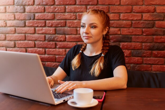 Young woman using laptop while sitting in cafe against brick wall