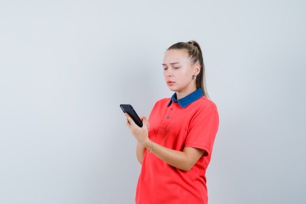 Молодая женщина с помощью мобильного телефона в футболке и обеспокоенным видом