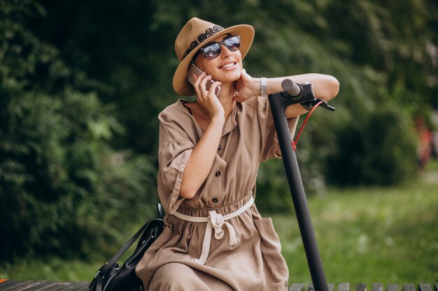 Young woman using phone in park sitting on bench by the scooter