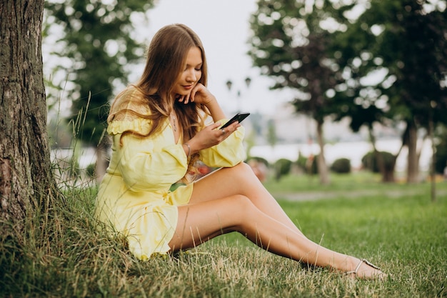 Giovane donna che utilizza il telefono e che si siede sotto l'albero nel parco