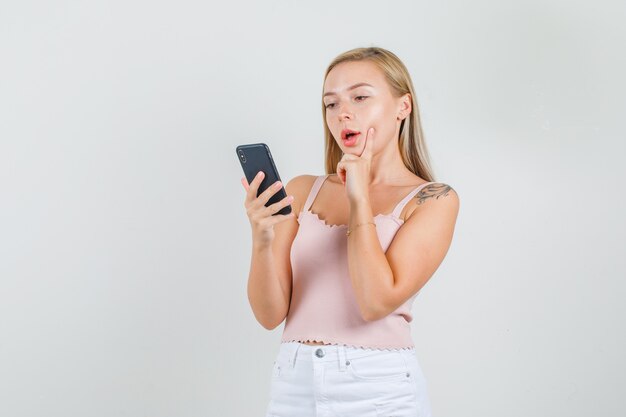 Young woman using smartphone with finger on cheek in singlet