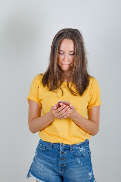 Young woman using smartphone in yellow t-shirt, shorts and looking perplexed
