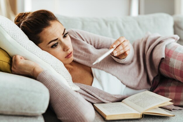 Young woman using thermometer and feeling worried while measuring her body temperature at home