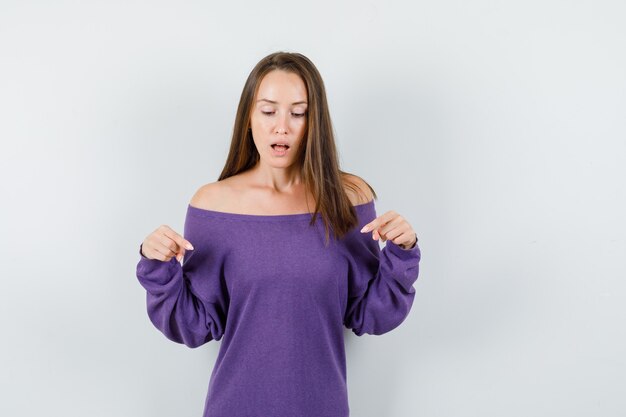 Young woman in violet shirt pointing down and looking focused , front view.