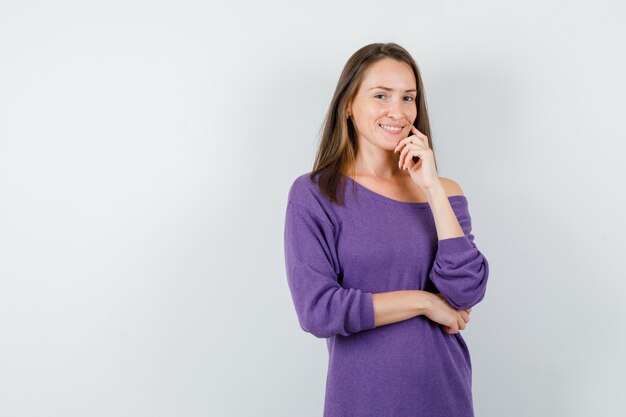 Молодая женщина в фиолетовой рубашке, стоящая в позе мышления и выглядящая веселой, вид спереди.
