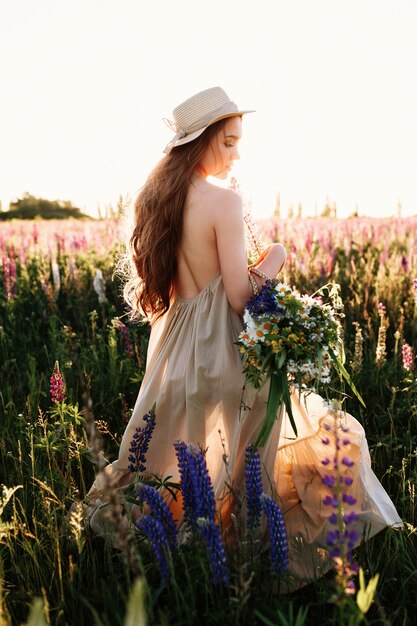 Young woman walking in flower field and high grass wearing hat and dress. 