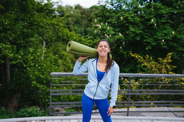 Молодая женщина, идущая в городском парке, держа коврик для фитнеса.