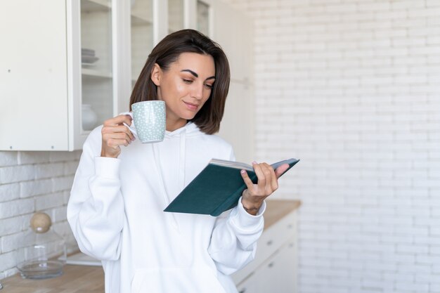 Young woman in a warm white hoodie at home in the kitchen reads a book in the morning