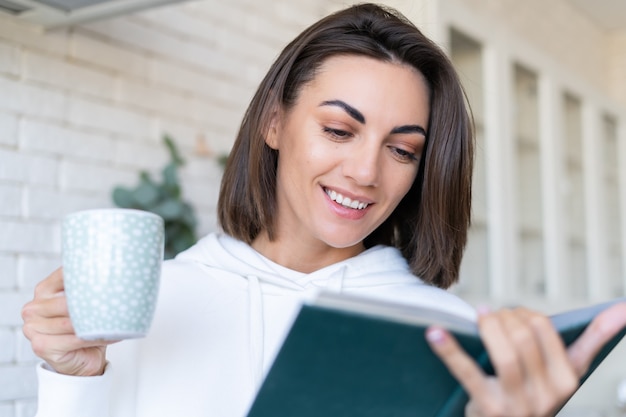 Young woman in a warm white hoodie at home in the kitchen reads a book in the morning