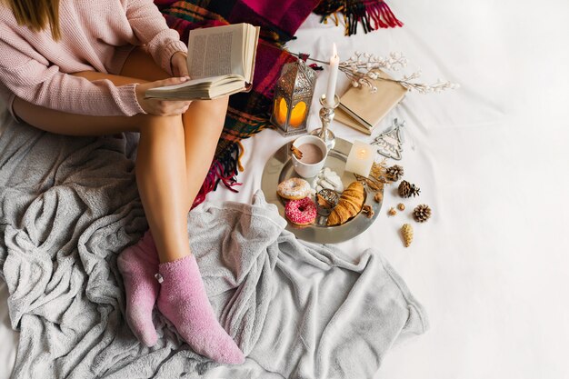 young woman in warm woolen clothes is sitting on her bed in bright sunny house