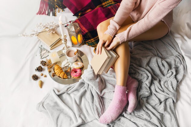 young woman in warm woolen clothes is sitting on her bed in bright sunny house