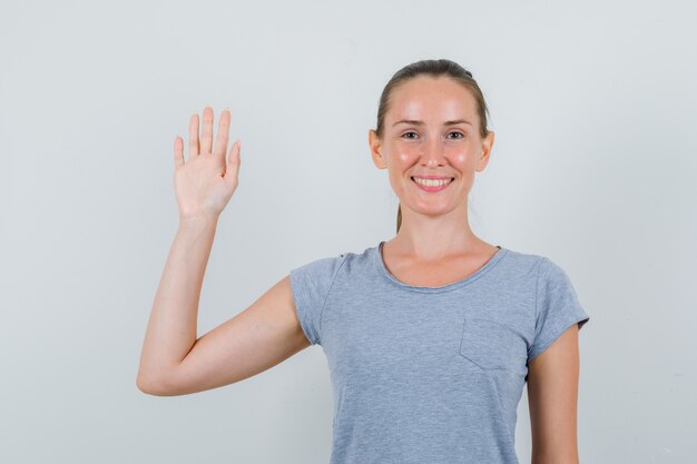 Молодая женщина машет рукой в серой футболке и выглядит веселой, вид спереди.