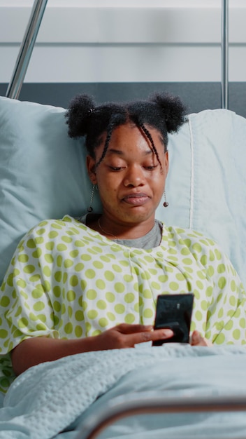 Young woman waving at video call camera on smartphone to talk to friends and family in bed. Patient with disease using online remote conference on mobile phone in hospital ward.