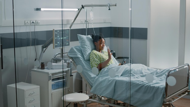 Young woman waving at video call on smartphone in hospital ward bed. Patient with IV drip bag and oximeter using online remote conference on internet to talk to friends and recovering