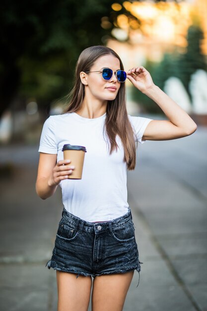 젊은 여자는 거리에 갈 커피와 함께 걷고 짧은, 흰 셔츠와 선글라스를 착용