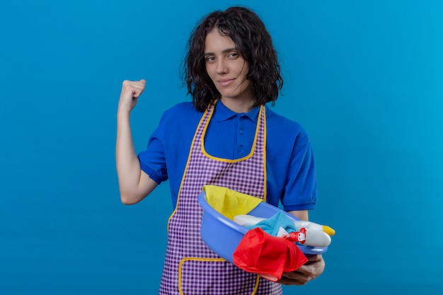 Free photo young woman wearing apron holding basin with cleaning tools raising fist showing biceps smiling confident, ready to clean over isolated blue wall