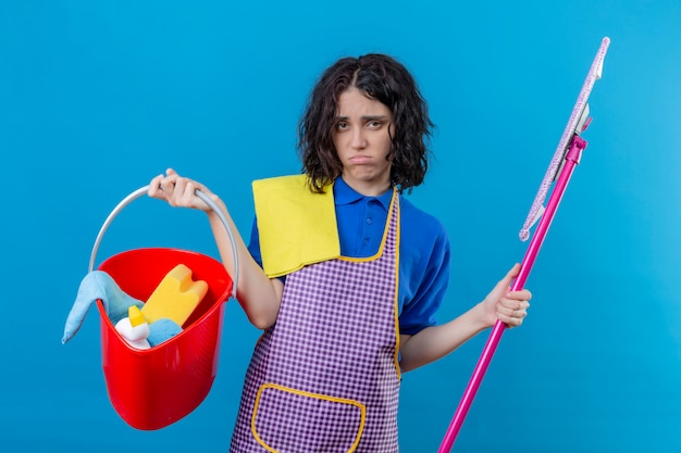 Foto gratuita secchio d'uso della tenuta del grembiule della giovane donna con gli strumenti e la zazzera di pulizia che sembrano sovraccarichi e stanchi soffiando le sue guance sopra la parete blu