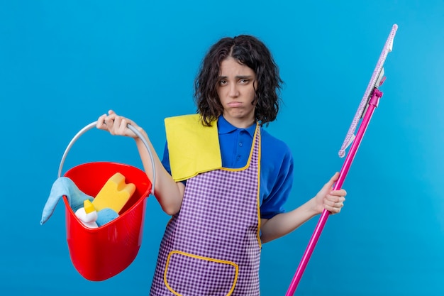 Foto gratuita giovane donna che indossa il grembiule tenendo la benna con strumenti di pulizia e mop cercando oberati di lavoro e stanchi che soffia le sue guance in piedi su sfondo blu