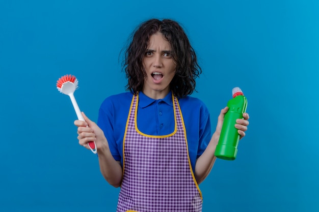 Foto gratuita la giovane donna che indossa il grembiule che tiene la spazzola e la bottiglia dei rifornimenti di pulizia ha colpito con la bocca spalancata sopra la parete blu