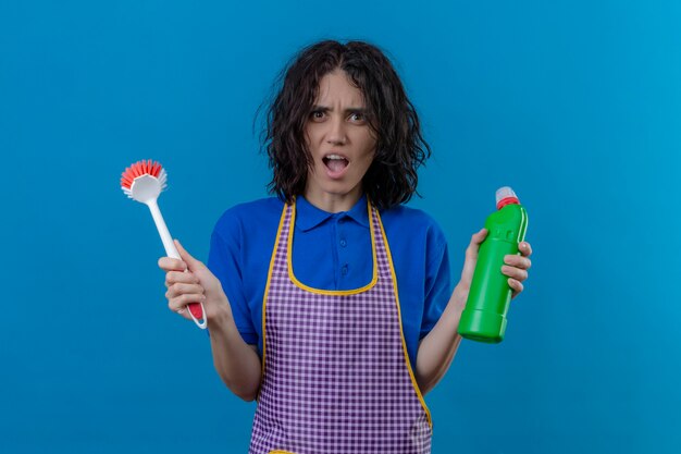 Young woman wearing apron holding scrubbing brush and bottle of cleaning supplies shocked with wide open mouth standing over blue background