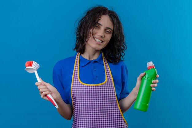 Young woman wearing apron holding scrubbing brush and bottle of cleaning supplies smiling cheerfully looking joyful over blue wall