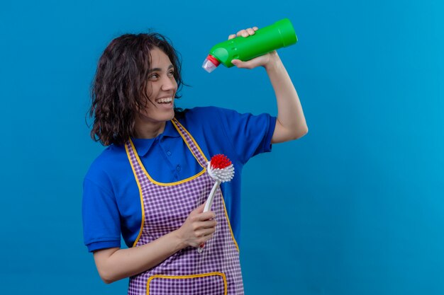 Young woman wearing apron holding scrubbing brush and bottle of cleaning supplies smiling cheerfully looking joyful over blue wall