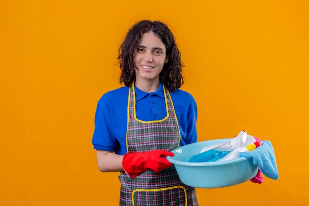 Young woman wearing apron and rubber gloves holding basin with cleaning tools looking confident smiling cheerful over orange wall