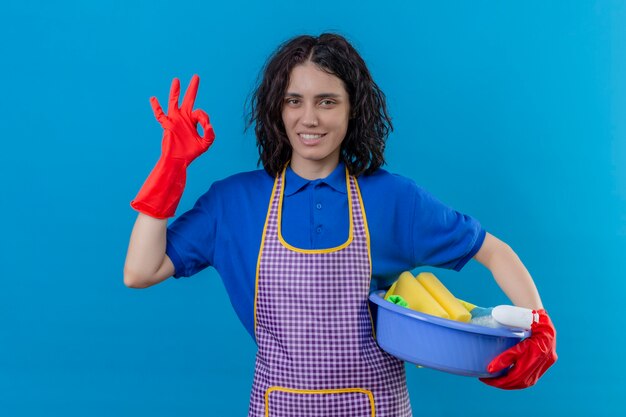 Молодая женщина в фартуке и резиновых перчатках, держащая таз с чистящими средствами, дружелюбно улыбается, делает хорошо, знак стоит на синем фоне