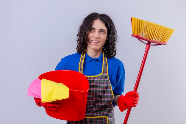 Young woman wearing apron and rubber gloves holding bucket with cleaning tools and mop looking up smiling and thinking over white wall