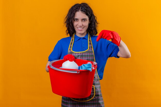 Young woman wearing apron and rubber gloves holding bucket with cleaning tools pointing with finger to it, smiling confident