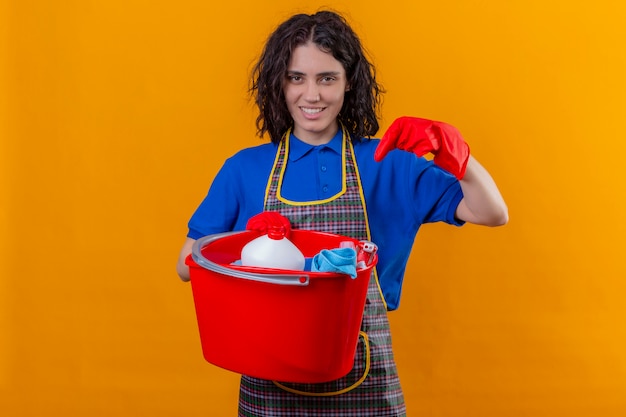 Young woman wearing apron and rubber gloves holding bucket with cleaning tools pointing with finger to it, smiling confident