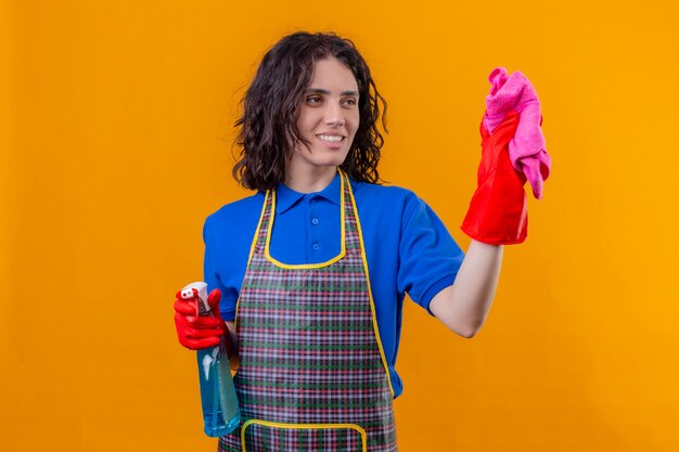 Young woman wearing apron and rubber gloves holding cleaning spray and rug smiling with happy face ready yo clean standing over isolated orange background