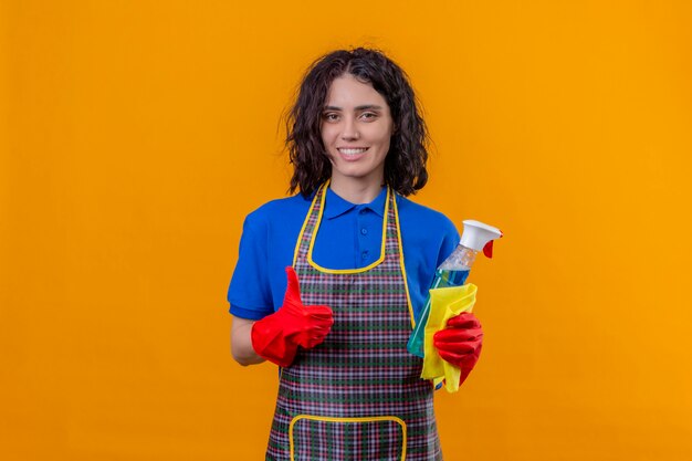 Молодая женщина в фартуке и резиновых перчатках держит чистящий спрей и коврик с большой улыбкой на лице показывает палец вверх над оранжевой стеной