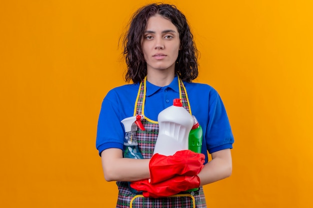 Grembiule d'uso della giovane donna e guanti di gomma che tengono i rifornimenti di pulizia con l'espressione sicura seria sopra la parete arancio isolata