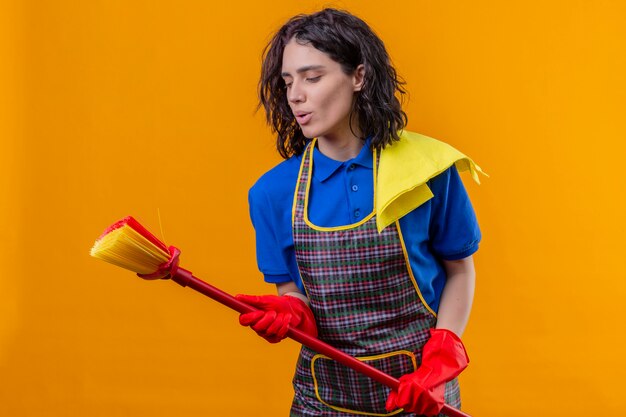 Young woman wearing apron and rubber gloves holding mop using as microphone singing a song, having fun over orange wall