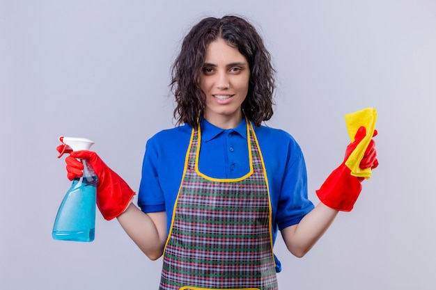 Young woman wearing apron and rubber gloves holding rug and cleaning spray with smile on face over white wall