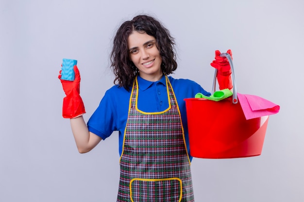 Foto gratuita grembiule d'uso della giovane donna e guanti di gomma che tengono spugna e secchio con gli strumenti di pulizia con il sorriso sul fronte sopra la parete bianca