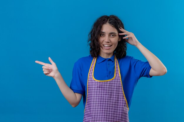 Foto gratuita grembiule da portare della giovane donna con il sorriso sul fronte e che indica il lato con il dito indice sopra la parete blu isolata