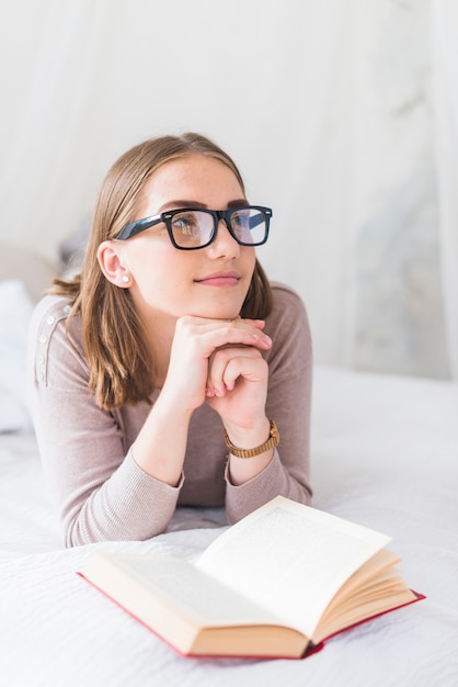 Young woman wearing black eyeglasses daydreaming while reading book on bed