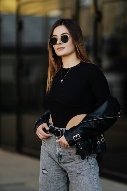 Young woman wearing black t-shirt, glasses and leather jacket posing against street