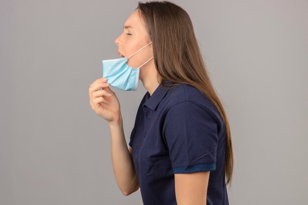 Young woman wearing blue polo shirt picking up mouth medical mask to cough feeling sick standing on light grey isolated background