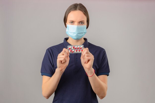 Молодая женщина в синей рубашке поло в защитной медицинской маске, держа в руках блистерные таблетки, смотрит в камеру с серьезным лицом на светло-сером фоне