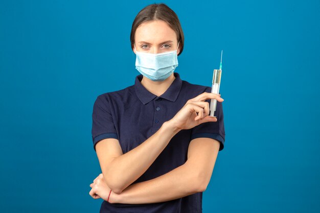 Young woman wearing blue polo shirt in protective medical mask holding syringe looking at camera with serious face standing on isolated blue background