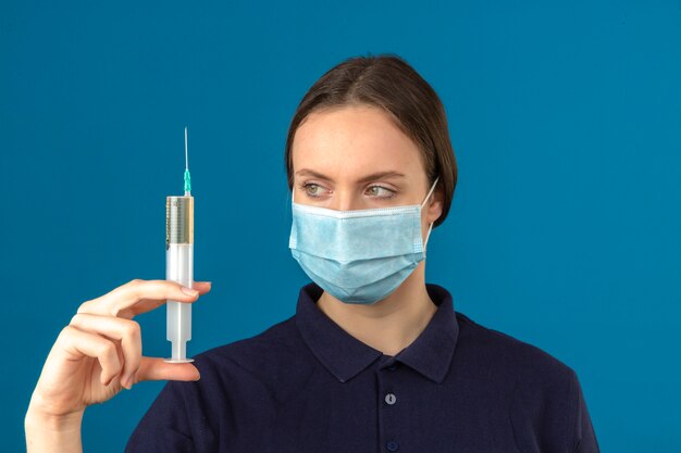 Young woman wearing blue polo shirt in protective medical mask holding syringe looking at it fixedly with serious face standing on isolated blue background