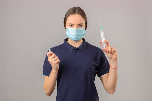 孤立した灰色の背景に深刻な顔立ちでカメラを見て注射器と薬のワクチンの瓶を保持している防護医療マスクで青いポロシャツを着た若い女性