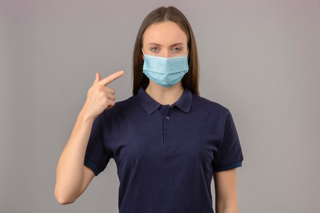 Young woman wearing blue polo shirt in protective medical mask pointing with finger on her mask with serious face looking at camera standing on light grey background